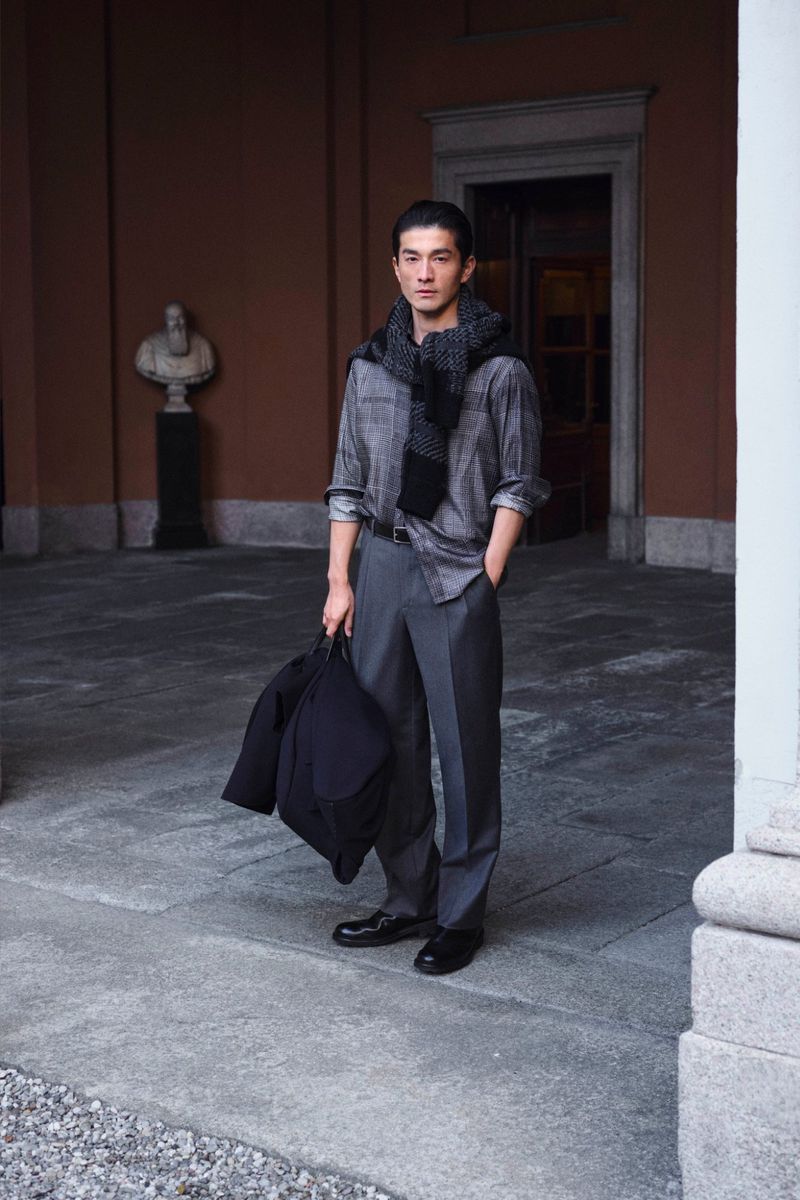 Full shot of an adult male in a grey shirt and trousers, holding a black jacket, standing outside an ornate building.