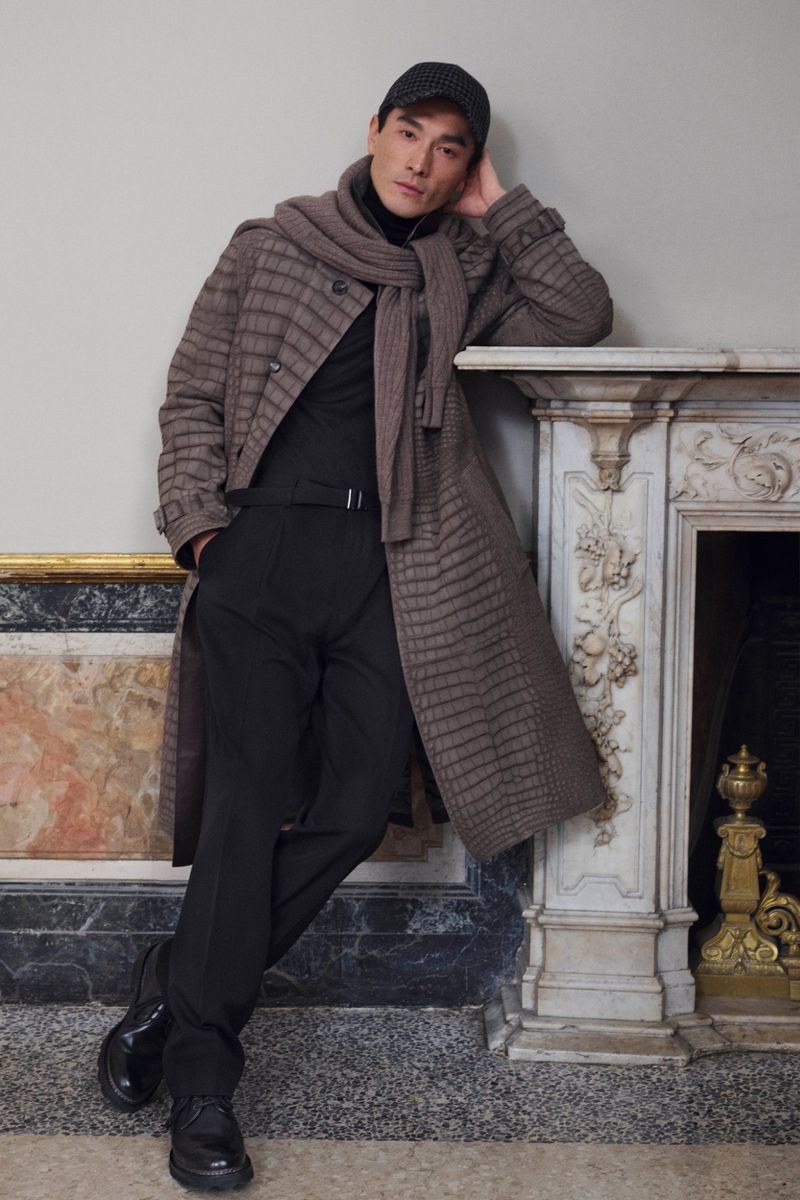 Stylish man in checked coat, scarf, and cap leans against marble fireplace, showcasing modern fashion in an indoor setting.