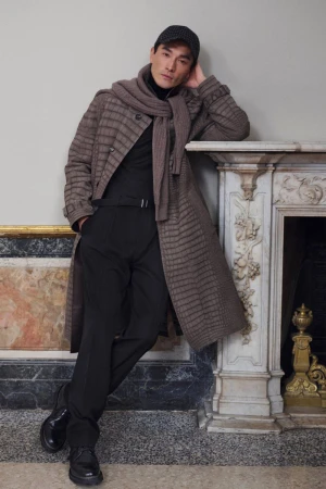 Stylish man in checked coat, scarf, and cap leans against marble fireplace, showcasing modern fashion in an indoor setting.