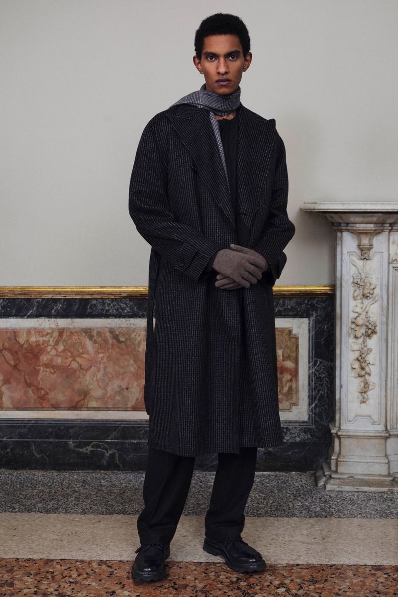 Man in gray overcoat and scarf poses indoors in front of a marble wall. He is wearing gloves, black pants, and black dress shoes.