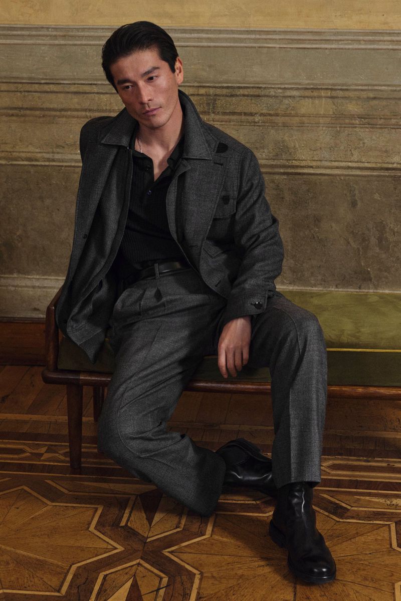 Asian man poses while seated, wearing a gray wool suit, black shirt, and boots in a stylish indoor fashion portrait.