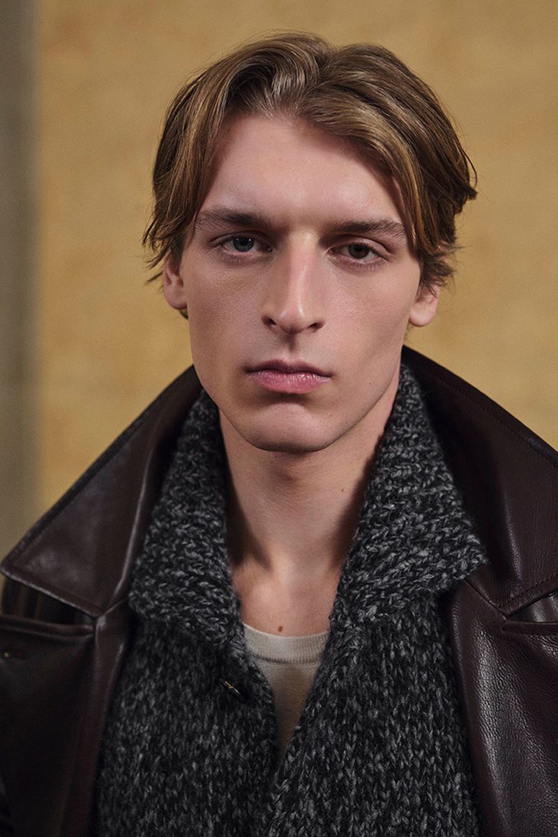 Close-up of a young adult male with blond hair wearing a brown leather jacket and grey knit sweater against a neutral background.