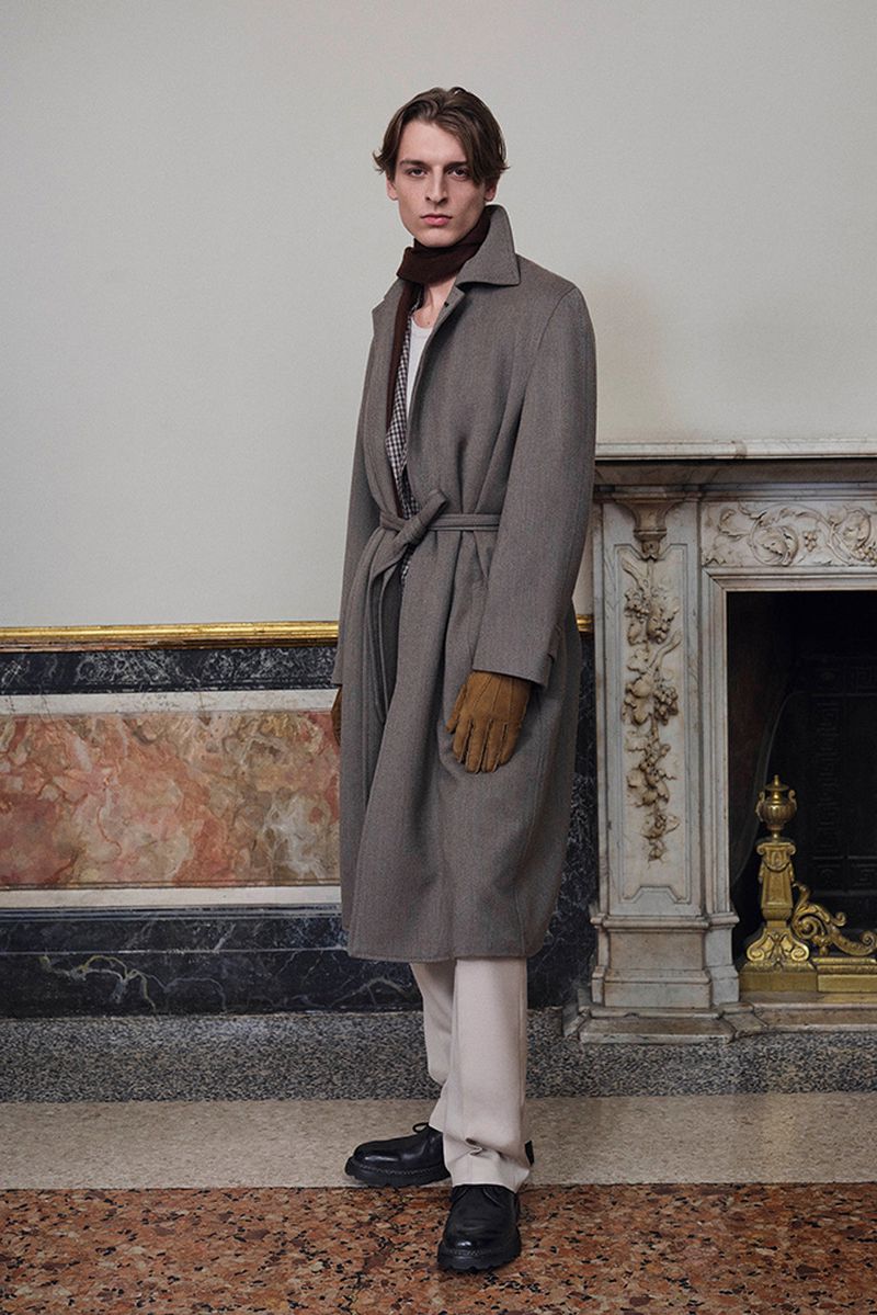 A stylish adult male stands indoors wearing a belted brown overcoat, trousers, scarf, gloves, and black shoes in a fashion setting.