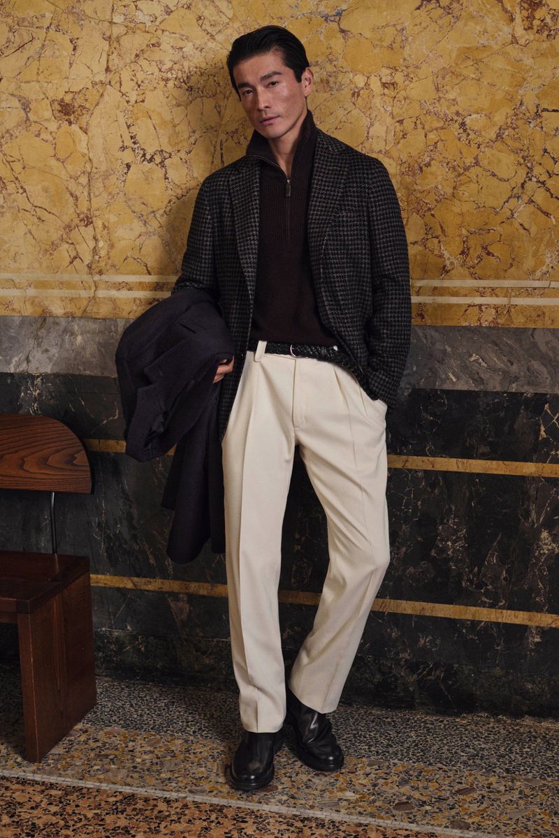 Man poses in a patterned overcoat, maroon sweater, and cream-colored dress pants indoors against a marble wall, holding a dark coat.