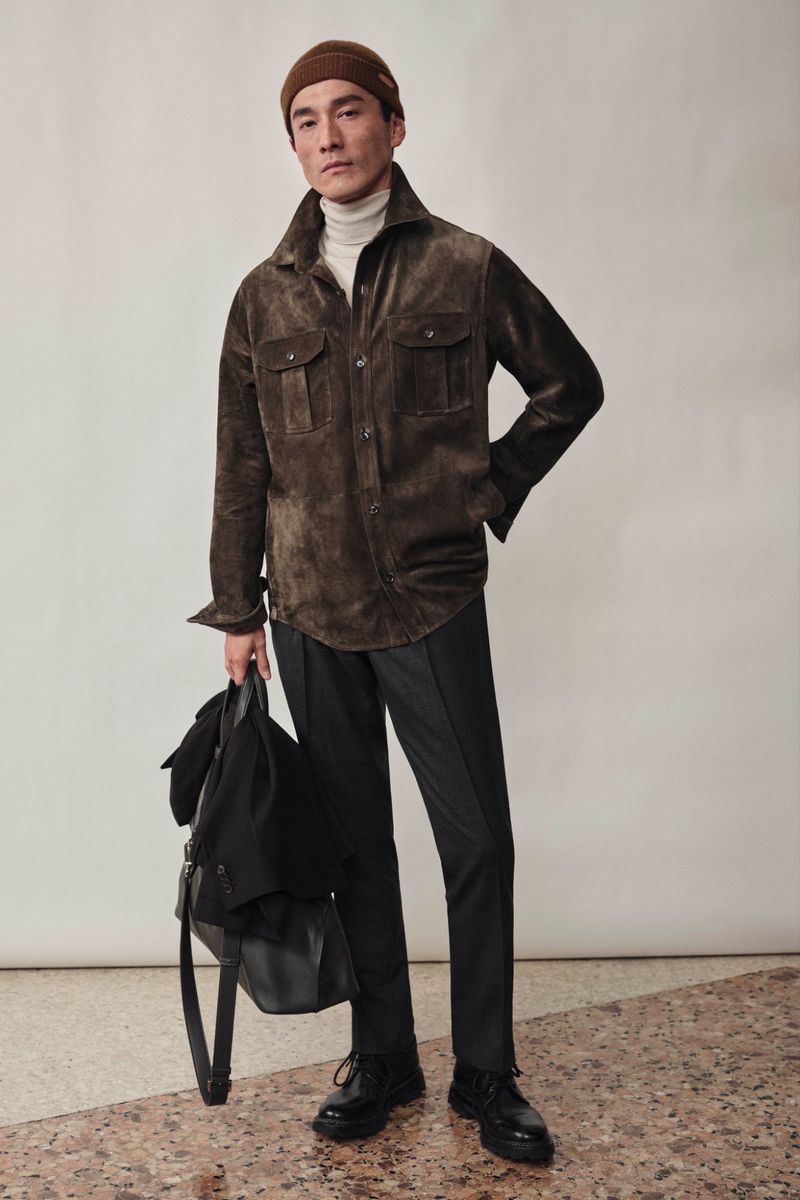 Asian male model in a brown suede jacket, beige turtleneck, and black pants stands in a studio, holding a black coat and bag.