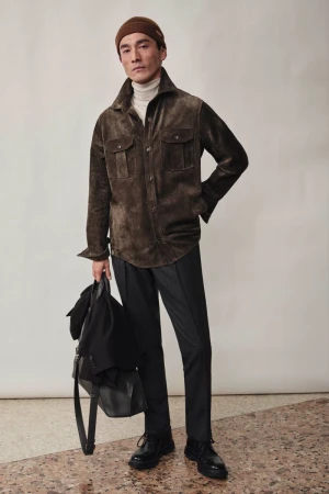Man wearing a brown leather jacket and beanie holding a black bag against a seamless studio backdrop