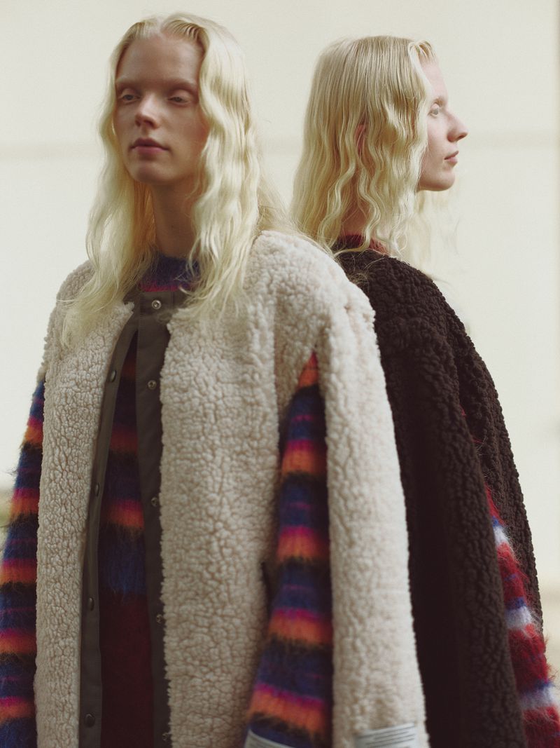 Two light-haired adults in a studio shot; one in a fleece jacket facing the camera, the other in a brown jacket in profile view.