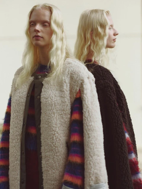 Two light-haired adults in a studio shot; one in a fleece jacket facing the camera, the other in a brown jacket in profile view.
