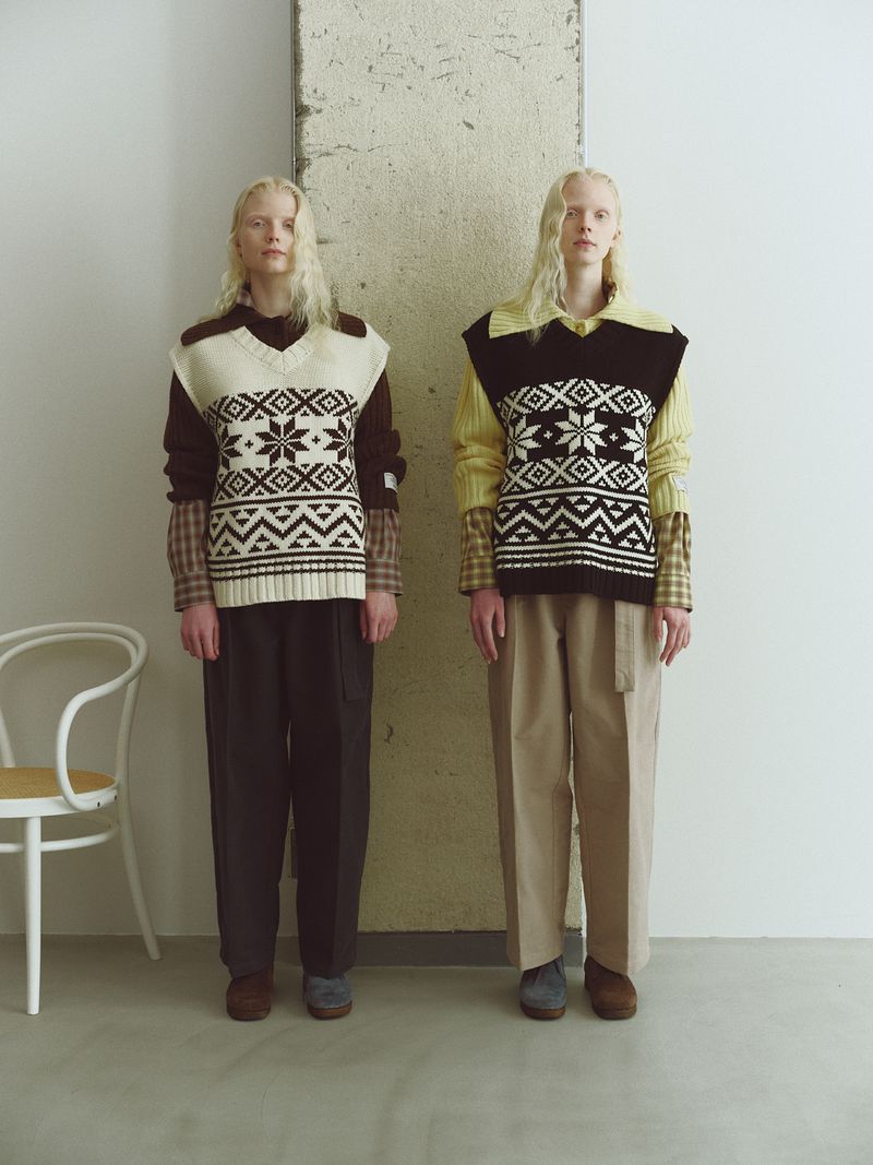 Two fair-skinned adult women pose indoors wearing patterned sweater vests, collared shirts, and trousers in a neutral color palette.