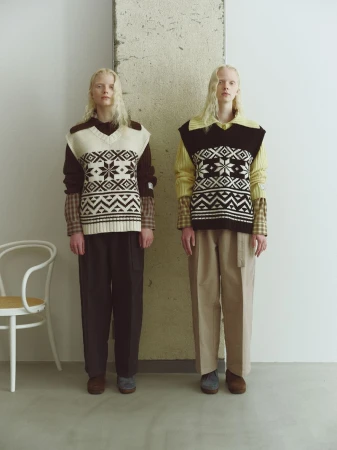 Two fair-skinned adult women pose indoors wearing patterned sweater vests, collared shirts, and trousers in a neutral color palette.