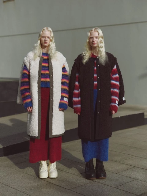 Two albino women stand side-by-side outdoors in colorful sweaters, midi skirts, and overcoats in front of a pale grey wall.