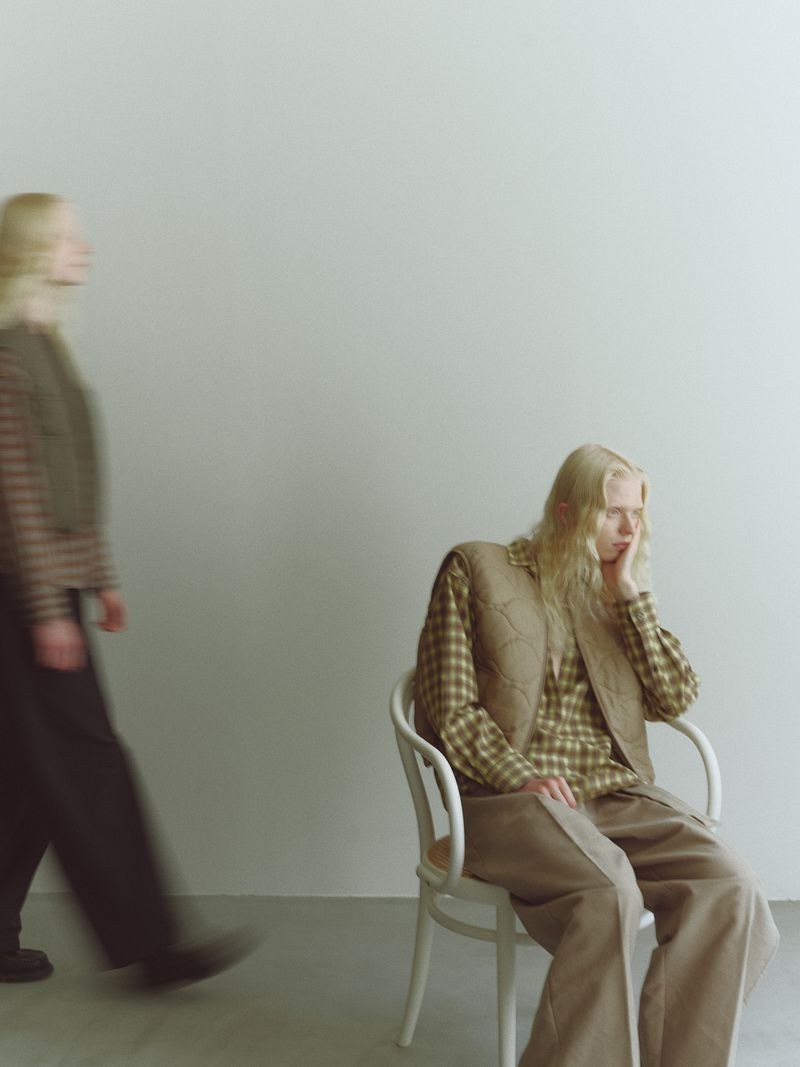 Two young women with blonde hair model fashion in a studio: one seated in a chair, the other walking in the background.