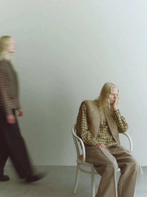 Two young women with blonde hair model fashion in a studio: one seated in a chair, the other walking in the background.