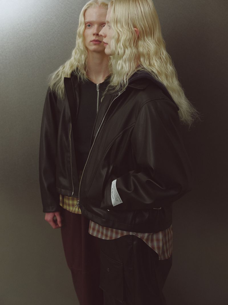 Two fair-skinned people with long blond wavy hair pose indoors wearing a black leather jacket, black hoodie, and plaid shirt.