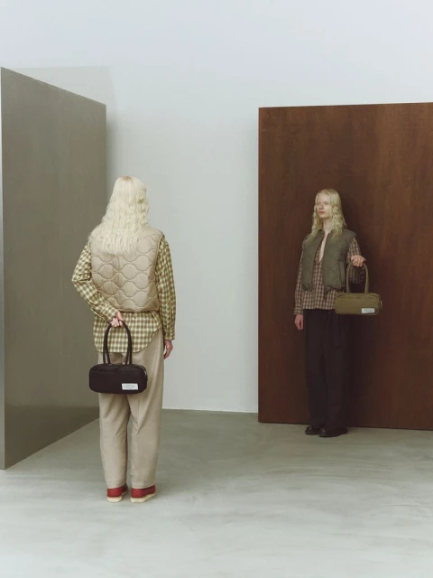 Two women modeling fashion handbags and vests in a photo studio with color blocks, wearing neutral-toned plaid shirts and pants.