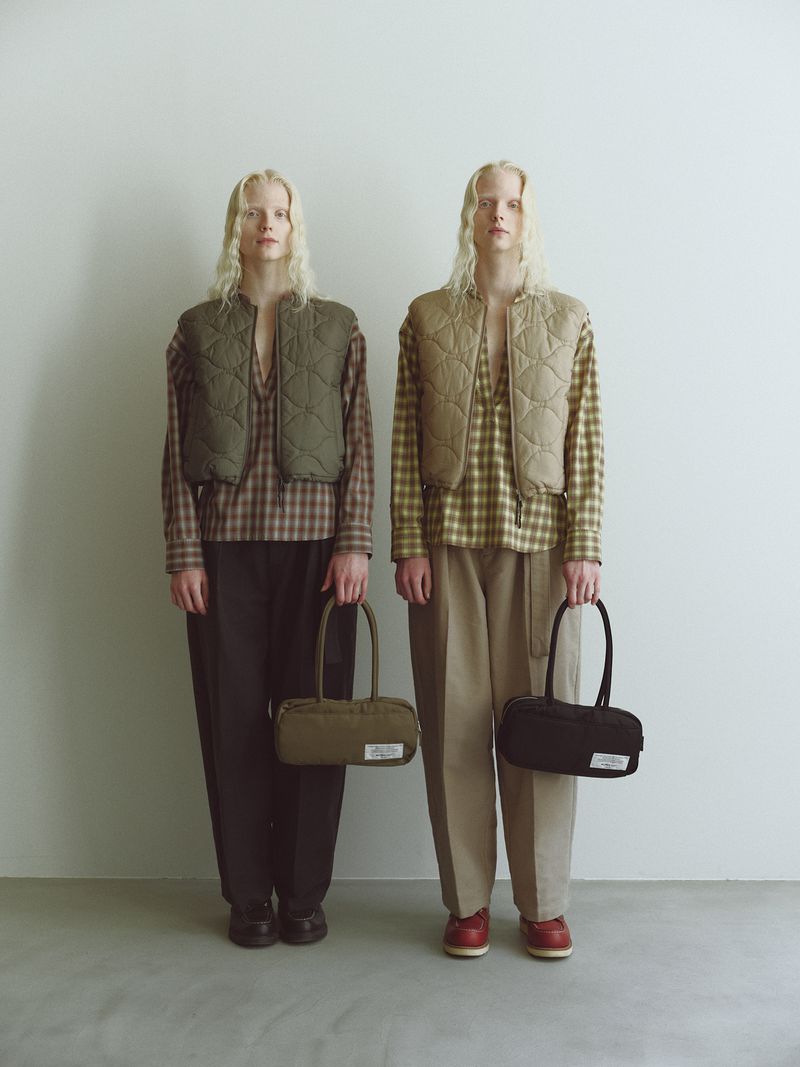 Two slender adult women pose in a studio setting, wearing cropped vests over plaid shirts, holding matching bags, fashionably coordinated.