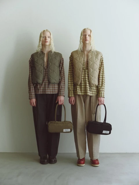 Two slender adult women pose in a studio setting, wearing cropped vests over plaid shirts, holding matching bags, fashionably coordinated.