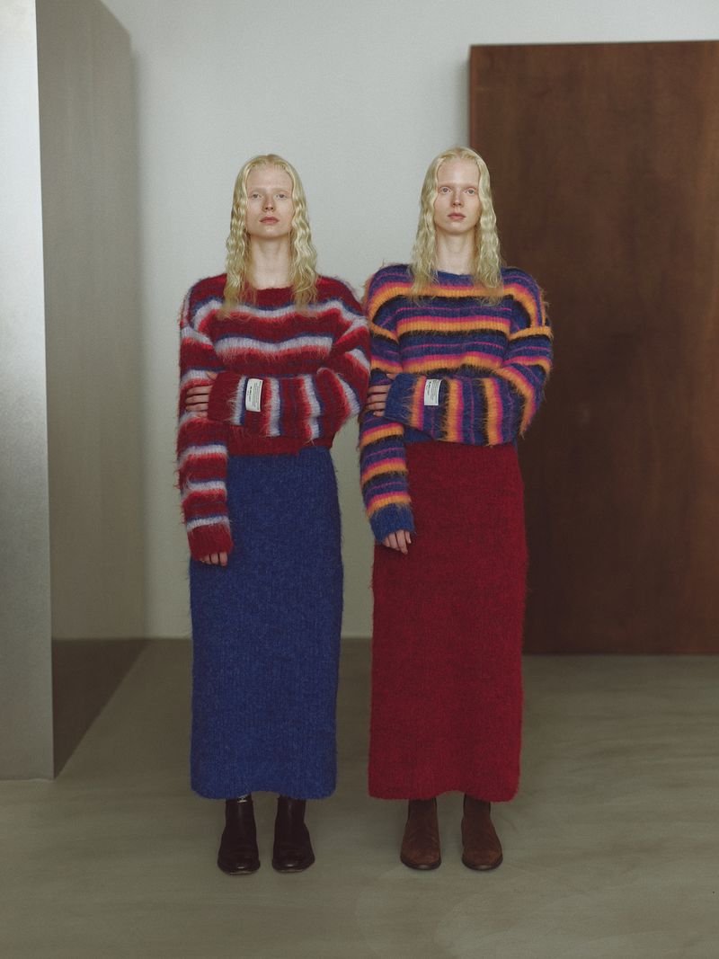 Two women wearing colourful striped sweaters and skirts stand indoors, their arms linked. Neutral colors and studio lighting enhance the fashion.