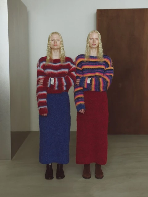 Two women wearing colourful striped sweaters and skirts stand indoors, their arms linked. Neutral colors and studio lighting enhance the fashion.