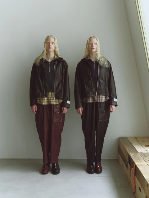 Two women in matching outfits: black bomber jackets, plaid shirts, and baggy pants. Dark footwear completes the coordinated look in a light-toned interior.
