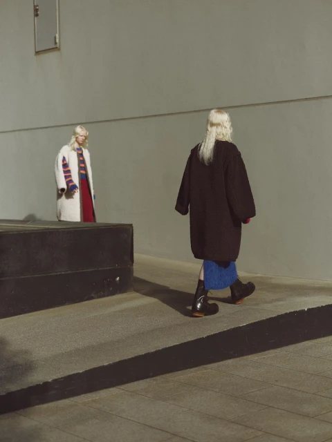 Two adult women in fashionable coats walking on an outdoor ramp, posing in front of a grey wall in urban daytime lighting.