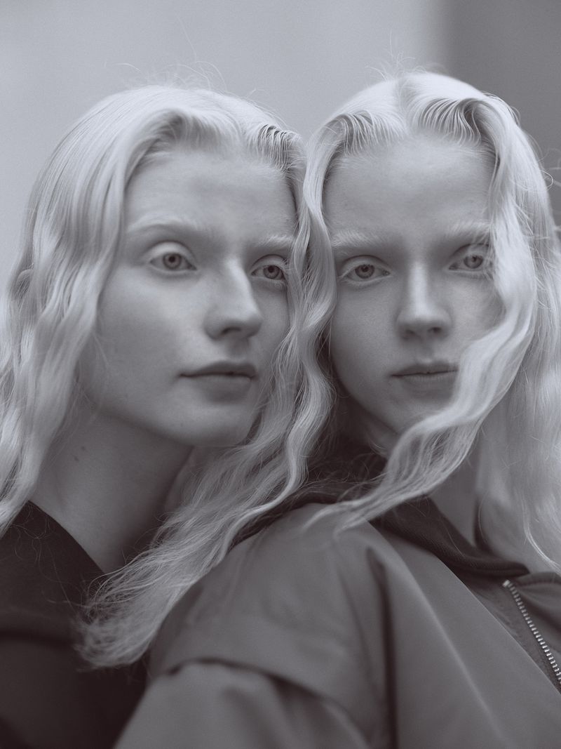 Monochrome portrait of two fair-skinned adult women with pale blonde hair, styled in a wavy texture, and matching jackets in a studio setting.