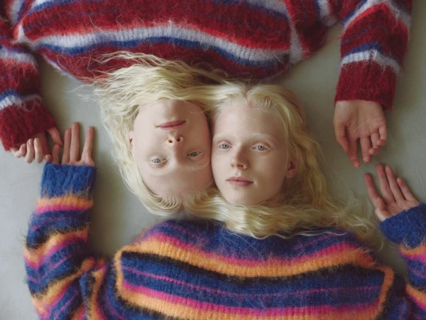 Two female models with long blonde hair lying head-to-head wearing colorful striped knit sweaters against a plain neutral background.