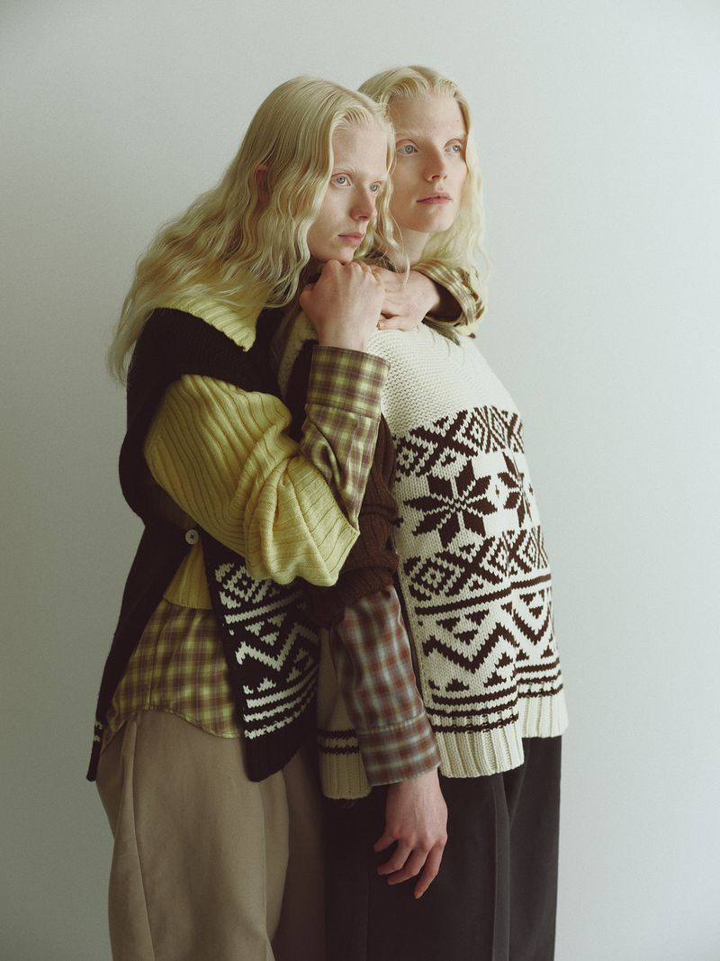 Two blonde models posing together wearing patterned knit sweaters against a plain white studio background.