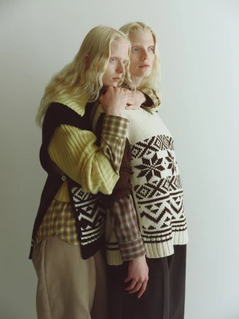 Two blonde models posing together wearing patterned knit sweaters against a plain white studio background.