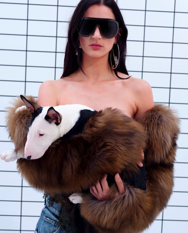 Woman with long dark hair wearing sunglasses and hoop earrings holds a bull terrier wrapped in fur against a white tile wall.