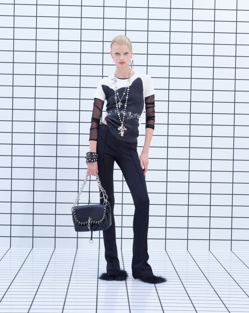 Model standing against a white tiled wall wearing black trousers and holding a black handbag for Blumarine