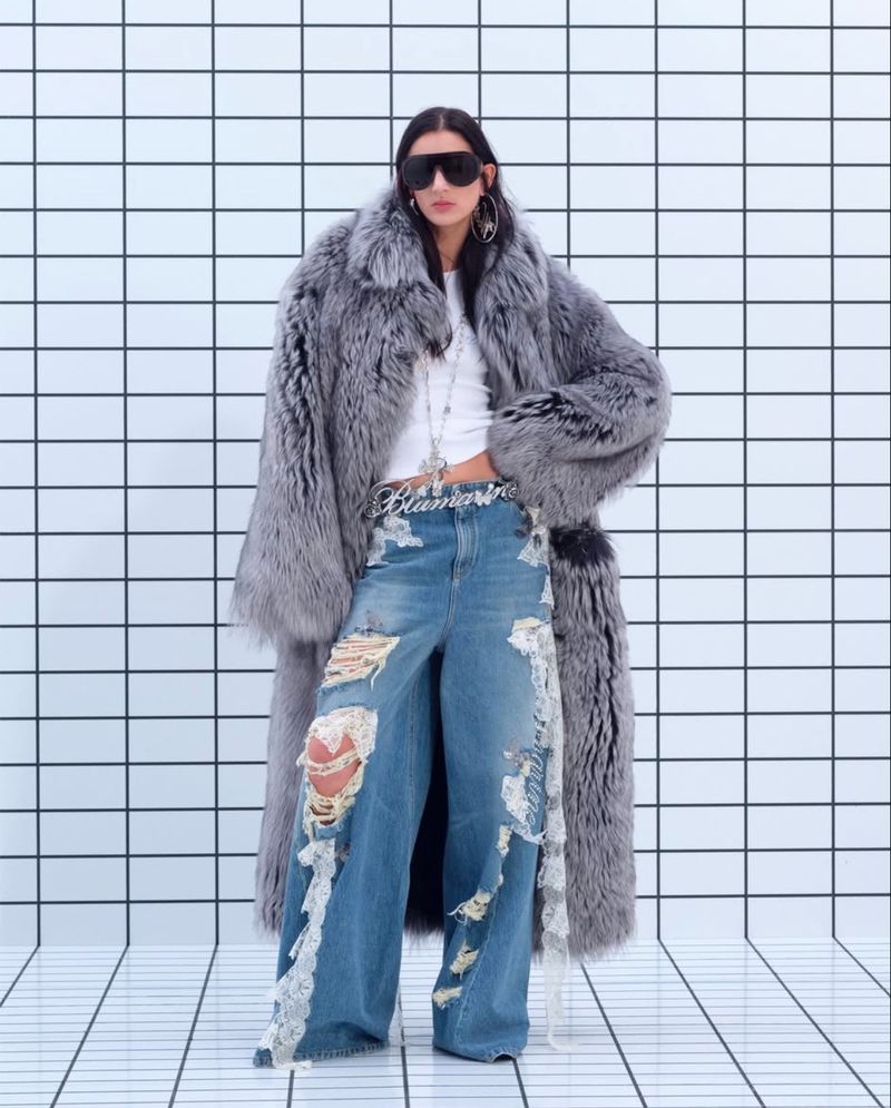 A woman in an oversized fur coat, distressed denim jeans, and sunglasses poses against a tiled wall in a fashion editorial shot.