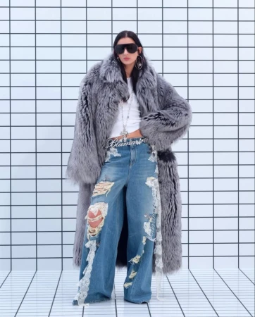 A woman in an oversized fur coat, distressed denim jeans, and sunglasses poses against a tiled wall in a fashion editorial shot.