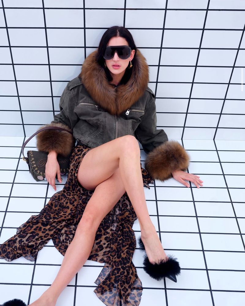 A woman in sunglasses poses in a fur-trimmed jacket and leopard print dress, seated on a tiled floor, with a purse nearby.