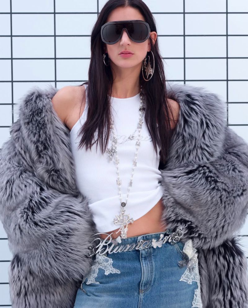 Model wearing dark sunglasses, grey fur coat, white tank top, and denim jeans against a tiled background