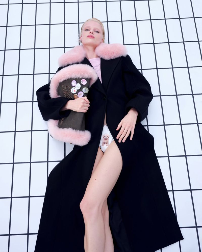 Woman in a black coat with pink fur trim lying on a white-tiled floor. She's holding a bag with buttons and wearing patterned shorts.