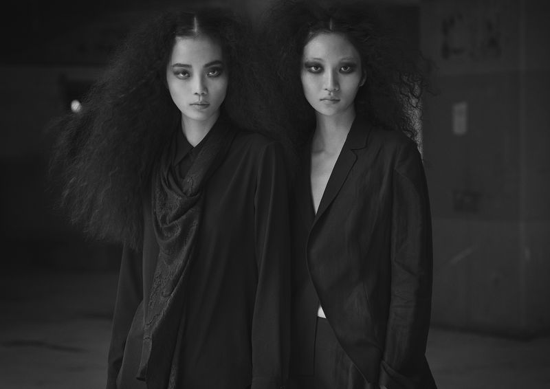 Black and white portrait of two Asian women with dark eye makeup and black, voluminous hair, wearing dark blouses in an indoor setting.
