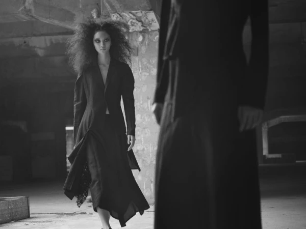 A woman in a dark dress and with curly hair stands inside of an empty building next to the vague silhouette of a person.