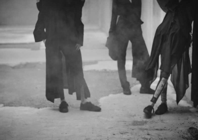Monochrome fashion campaign image featuring three models in dark layered clothing standing against a textured backdrop for Yohji Yamamoto.
