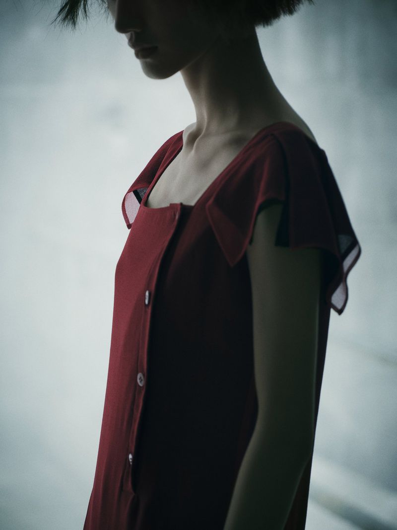 Portrait of a woman with short hair wearing a red dress with buttons and sheer flutter sleeves against a soft, diffused background.