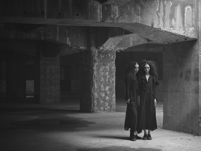Two women in dark coats stand in a stark, cavernous concrete building, lit by pale light from a high window, creating a somber mood.
