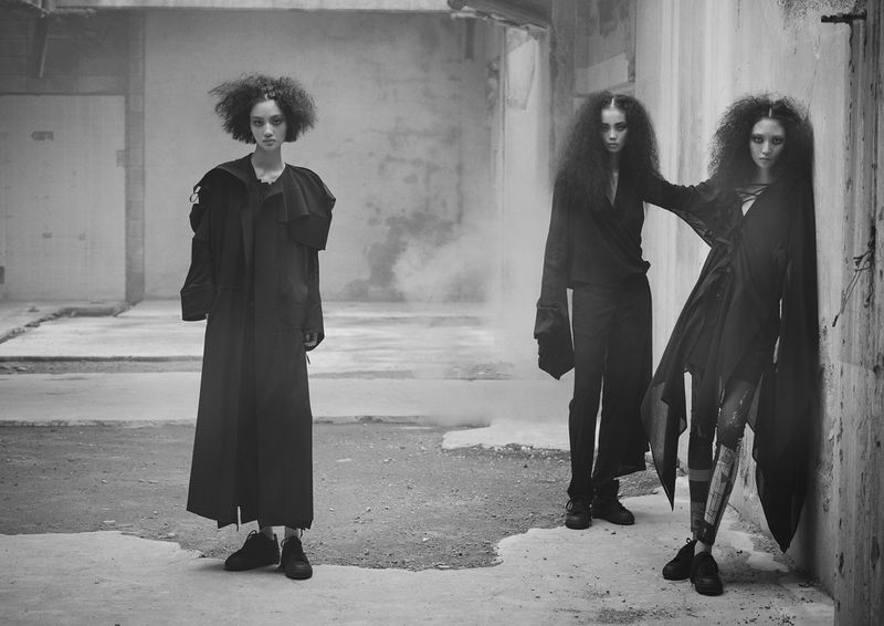 Three women pose in dark avant-garde fashion in an industrial area, captured in a monochromatic, medium shot with smokey, diffused lighting.