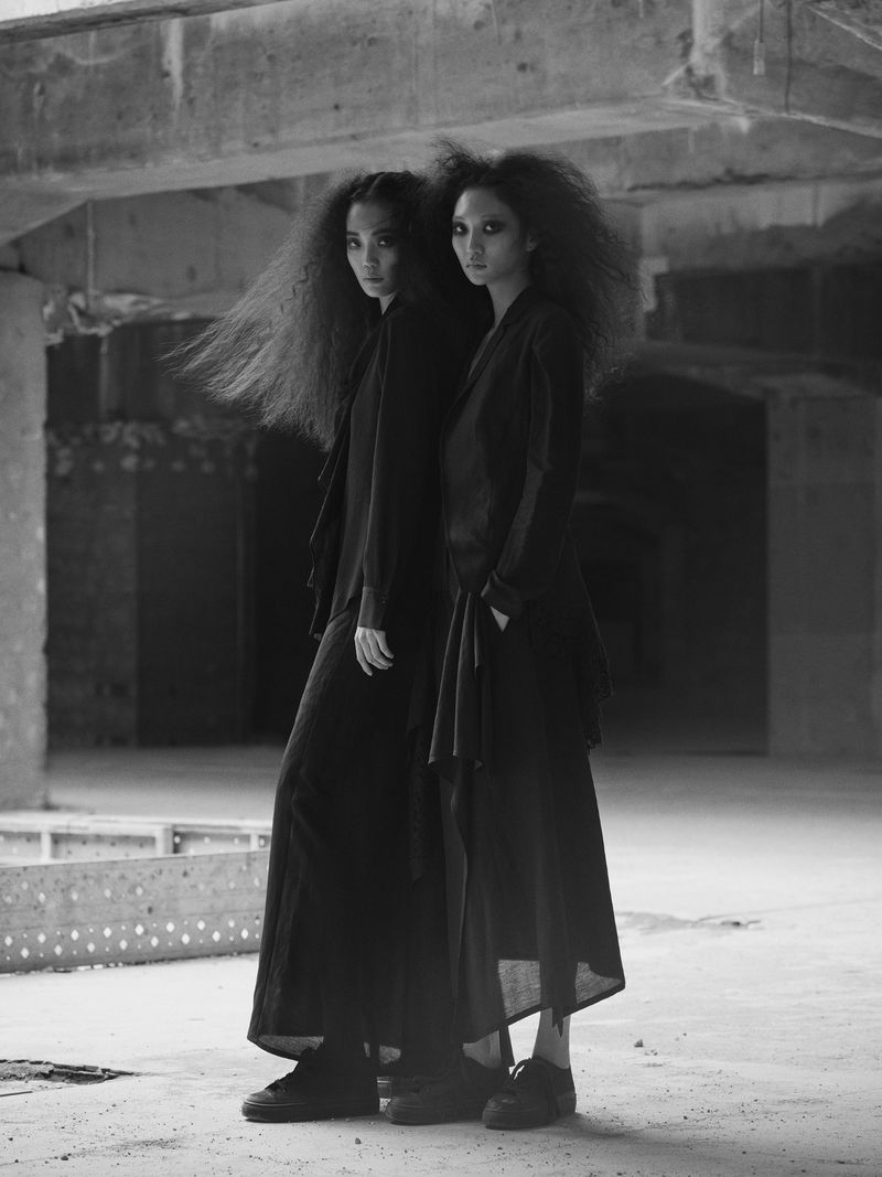 Two models in black outfits with voluminous textured hair standing in a concrete industrial interior