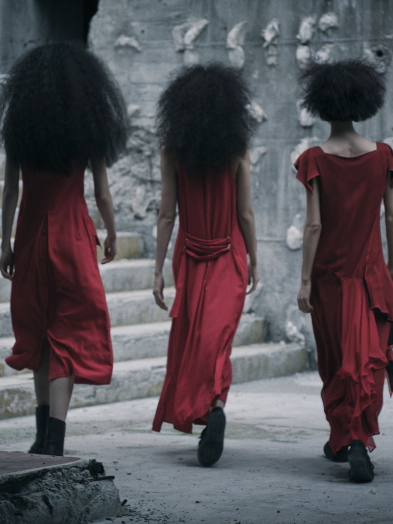 Three women in red dresses with large afro hairstyles walking away from the camera on a stone path in an outdoor setting.