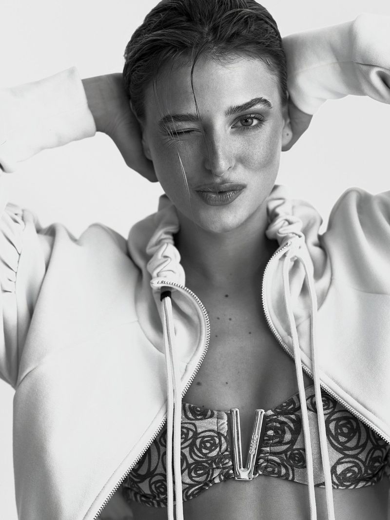 Monochrome portrait of a young adult female with short hair winking at the camera, wearing a floral top and unzipped hooded jacket.