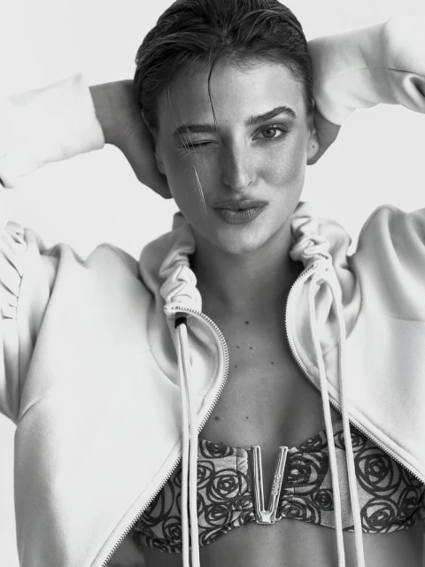 Monochrome portrait of a young adult female with short hair winking at the camera, wearing a floral top and unzipped hooded jacket.