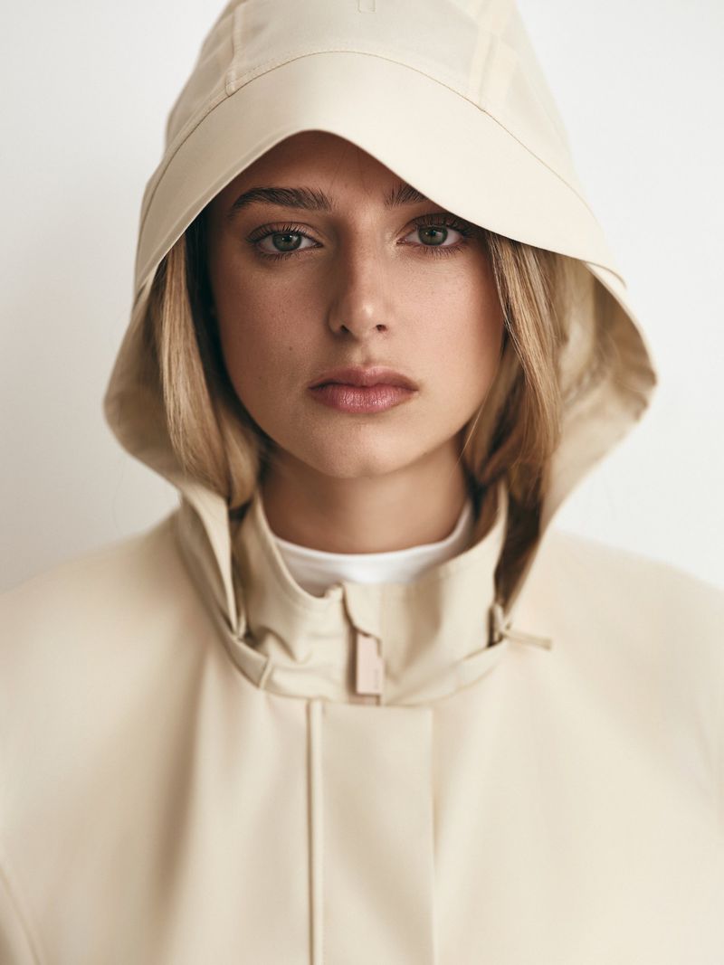 Close-up studio portrait of a blond woman in a beige hooded raincoat against a plain background, showcasing fashion and beauty.