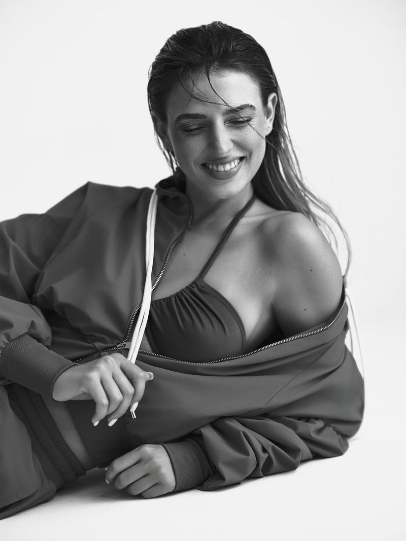 Smiling woman in a sports bra and jacket, lying down in a studio setting, black and white portrait with wet hair and natural light.