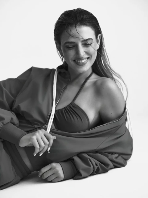 Smiling woman in a sports bra and jacket, lying down in a studio setting, black and white portrait with wet hair and natural light.