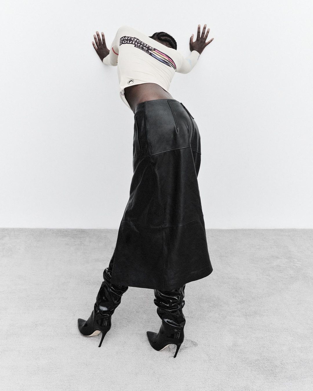 Fashion shot of a woman with dark hair in a white long-sleeve shirt, black leather skirt, and high-heeled boots against a white wall and floor.